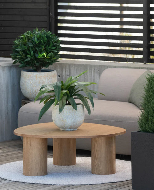 Sobre una pequeña alfombra junto a un sofá gris en una terraza hay una mesa de centro de madera con una planta artificial Bird's Deluxe de 60 cm encima, con unas cuantas plantas en macetas al fondo.