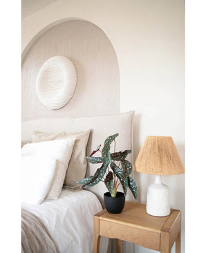 Un dormitorio acogedor con tonos neutros tiene una cama con ropa de cama blanca, una mesita de noche de madera con la Art Begonia de 48 cm en maceta decorativa negra y una lámpara tejida, y un arte de pared con textura redonda encima de la cama.