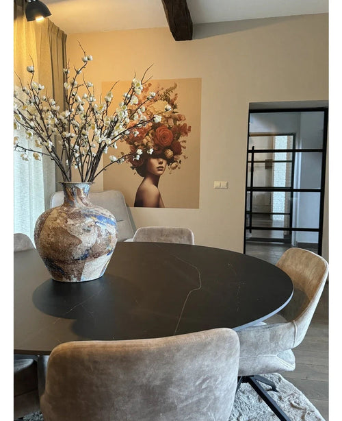 Un comedor moderno con una mesa redonda negra, sillas beige y un gran jarrón de cerámica con flor de cerezo japonés artificial de 94 cm en color blanco. Un mural de una mujer con flores en el pelo decora el fondo.