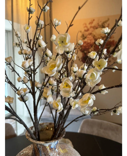 Un primer plano de la flor de cerezo japonés artificial de 94 cm con elegantes flores de cerezo artificiales blancas con centros amarillos sobre ramas marrones en un jarrón de cristal, sobre una mesa oscura con una habitación elegante y suavemente iluminada al fondo.