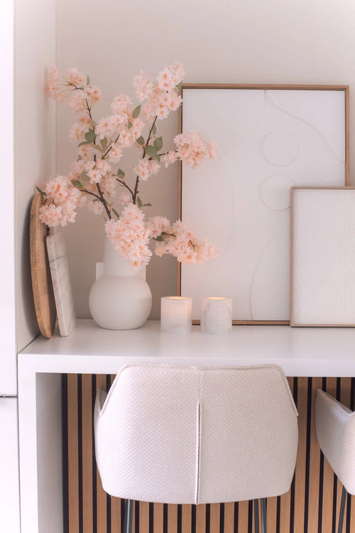 Un escritorio minimalista con una silla blanca, dos velas, bandejas decorativas, arte abstracto enmarcado y una flor de cerezo artificial de 100 cm de color rosa claro en un jarrón blanco sobre una superficie blanca.