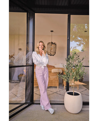 Una mujer con blusa blanca y pantalones color lavanda sonríe cerca de una gran puerta de cristal. El Olivo Artificial 120 cm aporta un ambiente mediterráneo al acogedor salón, con un sofá de mimbre y una decoración luminosa de fondo.