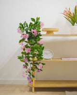 Una estantería de madera con planta colgante artificial Petunia de 70 cm violeta, un reloj blanco, un soporte de madera, un libro y un jarrón con flores rosas contra una pared blanca.