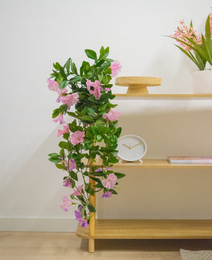 Una estantería de madera con planta colgante artificial Petunia de 70 cm violeta, un reloj blanco, un soporte de madera, un libro y un jarrón con flores rosas contra una pared blanca.
