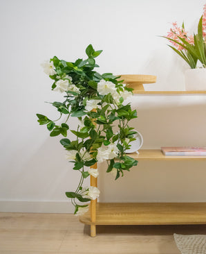 La planta colgante artificial Petunia 70 cm blanca con flores blancas cae de un estante de madera en una habitación luminosa y moderna con suelos de madera clara. En el estante superior hay un jarrón con largas flores rosas.