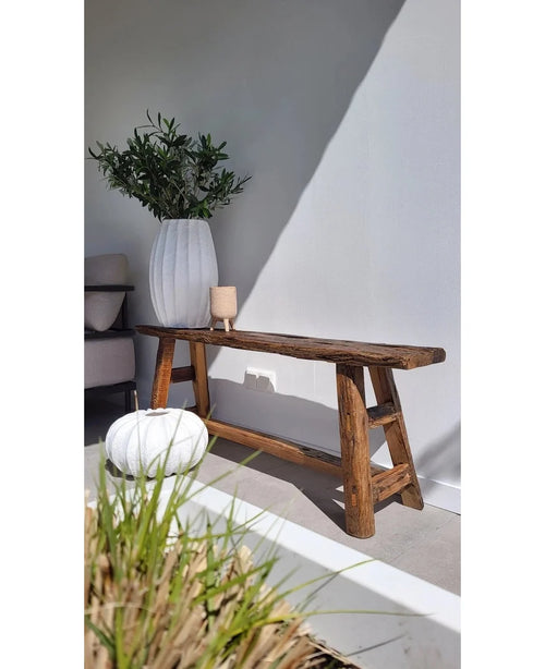 Un banco rústico de madera se apoya contra una pared blanca, junto a un jarrón blanco alto con hoja de olivo artificial de 90 cm UV para crear un ambiente mediterráneo. Cerca hay una otomana blanca redonda y un pequeño candelabro, con la luz del sol proyectando suaves sombras.