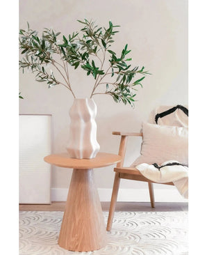 Un salón moderno con una silla de madera con un cojín luminoso, una mesa auxiliar redonda y un jarrón escultórico blanco con Hoja de Olivo Artificial 90 cm UV, creando un espacio minimalista con suaves tonos neutros y una sutil atmósfera mediterránea.