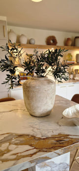 Un jarrón rústico beige con hoja de olivo artificial de 90 cm ignífugo se alza sobre una encimera de mármol en una cocina moderna y cálidamente iluminada, con estanterías y decoración mediterránea al fondo.