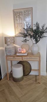 Una mesa consola de madera con dos cajones contiene una lámpara, un gran jarrón blanco con hoja de olivo artificial de 90 cm ignífuga, un jarrón más pequeño, libros y piedras decorativas. Los detalles mediterráneos y el arte abstracto enmarcado completan el look de arriba. En la parte inferior hay un taburete y una bandeja redonda.