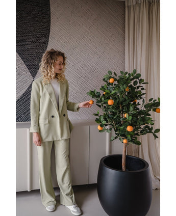 Een vrouw in een lichtgroen pak en witte sneakers staat binnen en raakt voorzichtig een oranje vrucht aan op de Kunst sinaasappelboom 120 cm naast haar, met een neutraal decor en een muur met patroon op de achtergrond.