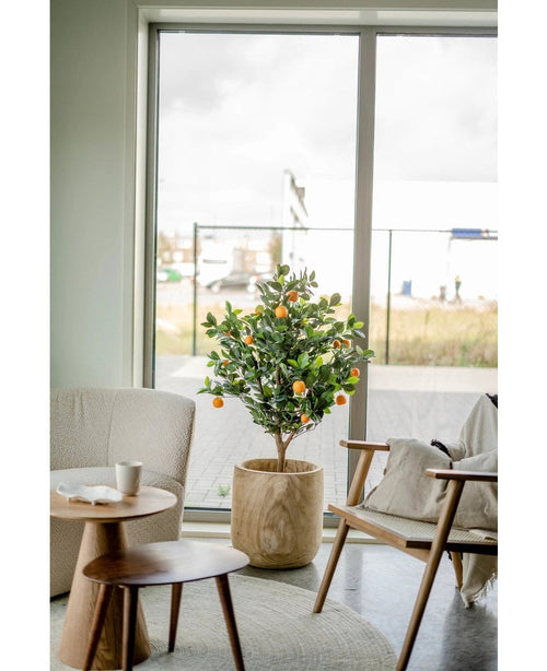 Un naranjo artificial de 120 cm se encuentra junto a un gran ventanal en un salón moderno y luminoso con una silla beige, una mesa auxiliar de madera y una taza de café sobre un platillo.