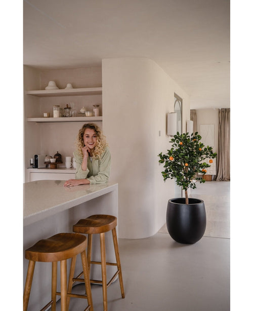 Una mujer de pelo rubio rizado sonríe, apoyada en un mostrador. Junto a ella, dos taburetes de madera y un naranjo artificial de 120 cm dan un ambiente fresco a la cocina moderna y luminosa, con estantes y platos detrás.