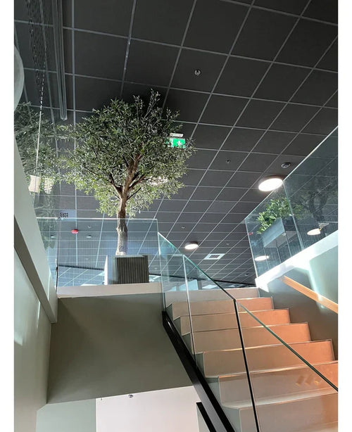 Una moderna escalera interior con barandillas de cristal conduce a un rellano que presenta el olivo artificial con aceitunas (270 cm) en una maceta grande, colocado bajo un techo con diseño de rejilla, iluminación empotrada y una señal verde visible de salida de emergencia.