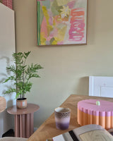 En una sala de temática pastel hay una planta de helecho artificial de 85 cm sobre una mesa auxiliar rosa, una taza morada y dorada, un cuenco rosa con el borde festoneado, un cuaderno abierto y en la pared un cuadro abstracto con tonos verdes, rosas y amarillos.