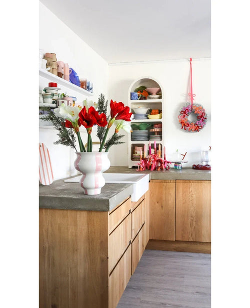 Una cocina luminosa con armarios de madera, estantes blancos abiertos con platos y ollas, una corona festiva en la pared y una flor artificial Amaryllis Real Touch roja de 36 cm sobre la encimera de hormigón.