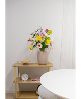 Een lichthouten plank toont een beige vaas met Kunstranonkel Real Touch Geel 60cm bloemen, een witte klok en een houten dienblad. Een deel van een ronde witte tafel verschijnt op de voorgrond tegen een effen witte muur.