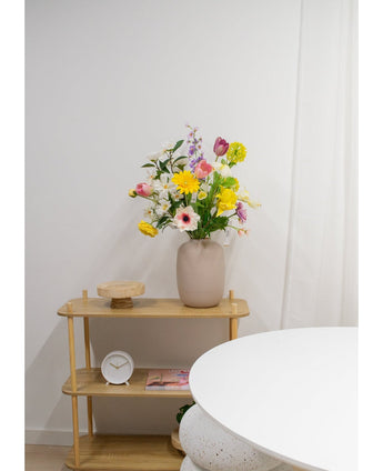 Un estante de madera clara muestra un jarrón beige con flores artificiales Ranunculus Real Touch Yellow de 60 cm, un reloj blanco y una bandeja de madera. Parte de una mesa redonda blanca aparece en primer plano contra una pared blanca y lisa.