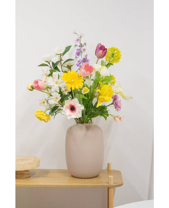 Een beige vaas met een Kunstranonkel Real Touch Geel 60cm kunstbloem staat op een licht houten tafeltje tegen een effen witte muur.