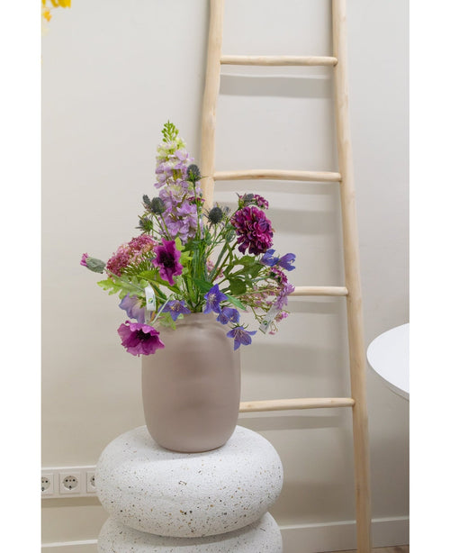 Una flor artificial de campanilla de 62 cm de color violeta se encuentra en un jarrón de vidrio esmerilado sobre una base blanca estructurada. Detrás, una escalera de madera clara está apoyada contra una pared beige, con enchufes visibles cerca del suelo.