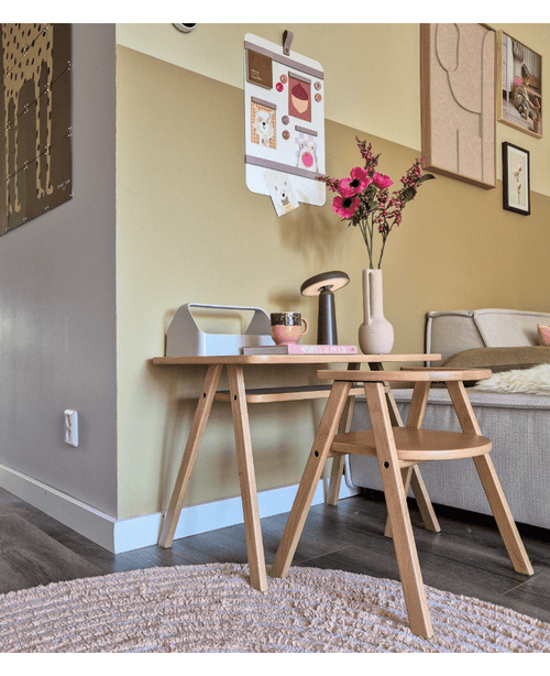 Un rincón de salón moderno con una mesa de centro de madera clara con ramas de flores artificiales de anémona de 56 cm de color rosa en un jarrón rosa, junto con una lámpara y libros. Al lado hay un sofá beige, acentuado por arte en la pared y un tablero de fotografías tipo galería en la pared amarilla.
