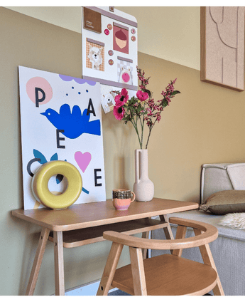 Een houten bureau met een bijpassende stoel toont een blauwe vogelafdruk, Kunstbloem Anemoon vertakt 56 cm roze, een rond geel ornament en een klein kopje. Boven hangt een prikbord met foto's en notities naast een beige bank.