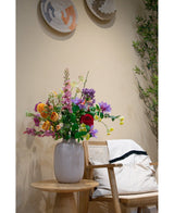 Un jarrón blanco con Cornejo Artificial Real Touch Amarillo Claro de 74 cm se encuentra sobre una mesa auxiliar de madera junto a una silla de madera clara con un cojín blanco y una alfombra con bordes negros, acentuada por una decoración de pared tejida y una planta verde en el fondo.
