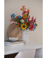 Un ramo de flores de clavel artificial de 70 cm de color azul en un jarrón de cerámica blanca se encuentra sobre un escritorio blanco junto a una pila de libros y un pequeño cuenco decorativo, con una bandeja redonda de madera apoyada contra la pared detrás.