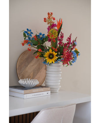 Un ramo de flores de clavel artificial de 70 cm de color azul en un jarrón de cerámica blanca se encuentra sobre un escritorio blanco junto a una pila de libros y un pequeño cuenco decorativo, con una bandeja redonda de madera apoyada contra la pared detrás.