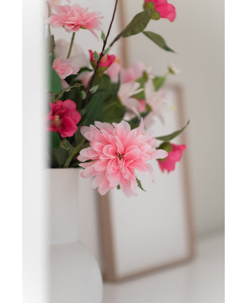Un primer plano de la flor artificial Dalia de 68 cm rosa en un jarrón blanco, con un tenue marco de fotos al fondo sobre una superficie blanca.