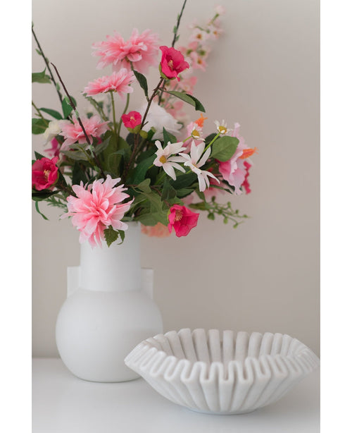 Una flor artificial Dalia de 68 cm de color rosa en un jarrón blanco se encuentra junto a un cuenco de cerámica blanca con volantes sobre una superficie blanca, sobre un fondo liso y claro.
