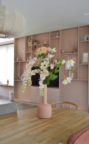 Un comedor moderno con una mesa de madera con un jarrón color melocotón que contiene la Flor Artificial Fritillary 94 cm blanca. Al fondo hay una gran estantería empotrada decorada con elementos elegantes.