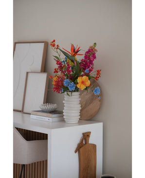 Un jarrón blanco acanalado con flor artificial de hierba gatera de 75 cm rosa se encuentra sobre un escritorio blanco con libros, arte enmarcado, una bandeja de madera y una tabla de cortar, junto a una silla beige en una habitación moderna y minimalista.