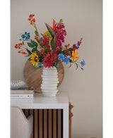 Un jarrón blanco texturizado sostiene un ramo de flores artificiales de hierba gatera rosa de 75 cm sobre una mesa blanca junto a libros apilados y decoración, con una bandeja redonda de madera apoyada contra la pared beige al fondo.