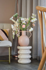 La flor artificial Fritillary de 94 cm blanca se encuentra en un jarrón de cerámica rosa claro con asas, colocada sobre una base blanca ondulada junto a una silla moderna con un cojín a rayas y cortinas neutras en una habitación elegante.