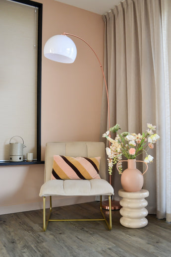 Un rincón moderno y acogedor con una silla color crema con un cojín a rayas, una lámpara de pie alta en forma de arco, cortinas beige, suelo de madera y un jarrón redondo con flores blancas Artificial Flower Fritillary de 94 cm sobre una mesa auxiliar esculpida.