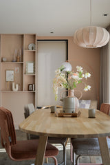 Een moderne eetkamer met een lichthouten tafel, vier stoelen met kussens, de Kunstbloem Fritillary 94 cm wit in een vaas, keramische kopjes, pastelroze muren, ingebouwde planken met decor en een grote ronde hanglamp boven je hoofd.