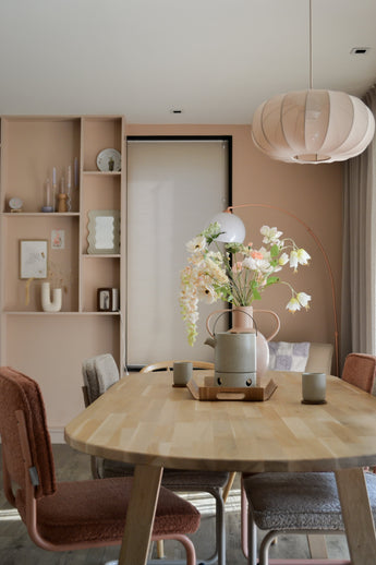 Un comedor moderno con una mesa de madera clara, cuatro sillas con cojines, la Flor Artificial Fritillary de 94 cm blanca en un jarrón, tazas de cerámica, paredes de color rosa pastel, estantes empotrados con decoración y una gran lámpara colgante redonda sobre tu cabeza.