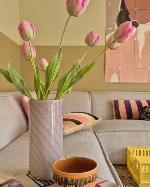 Una flor artificial de tulipán francés de 63 cm en color rosa se encuentra en un jarrón sobre la mesa junto a una taza naranja en una acogedora sala de estar con un sofá color crema, cojines estampados, una caja amarilla y arte abstracto en la pared al fondo.
