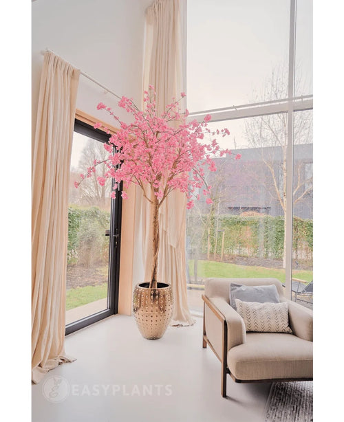 El árbol de cerezo rosa artificial de 240 cm se encuentra en una maceta decorativa junto a un gran ventanal en un salón luminoso y moderno con cortinas de color crema, un sillón beige y vistas al jardín exterior.