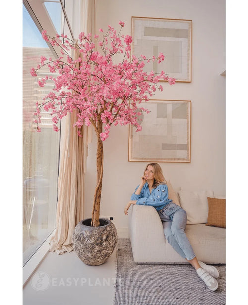 Una mujer vestida de forma informal se relaja en un sofá beige junto a un árbol de cerezo artificial de 240 cm con flores rosadas, colocado en una sala de estar moderna con arte abstracto en las paredes y grandes ventanales.