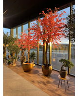 Potplanten en -bomen, zoals de Kunstboom Kersenbloesem roze van 330 cm met felroze bloesems, staan opgesteld bij hoge glazen ramen die het licht op de houten vloer weerkaatsen.