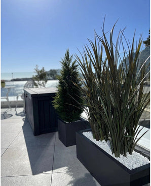 Césped artificial Burdeos UV 120 cm en jardineras rectangulares con piedras blancas decora una moderna terraza exterior con un fondo de cielo azul y árboles a lo lejos.