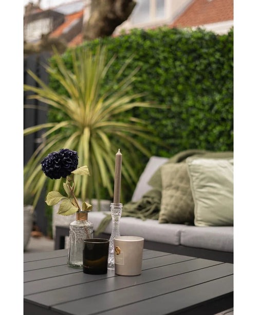 Una moderna mesa de exterior con superficie negra muestra un jarrón con una flor oscura, un candelabro de cristal y dos tazas. Detrás hay un sofá con cojines verdes frente a una pared verde exuberante hecha con la mezcla de alfombras Kunsthaag de 50x50 cm.