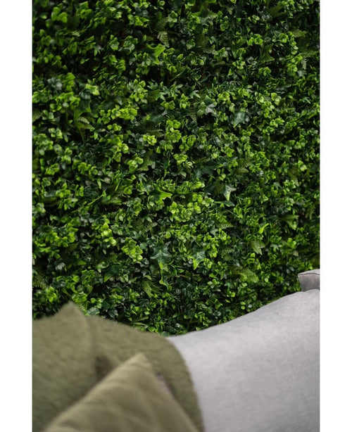 Primer plano de un seto artificial de color verde exuberante que mezcla una pared de 50 x 50 cm con hojas vibrantes, con la esquina de un sofá gris claro y parte de un abrigo verde visible en el primer plano inferior.