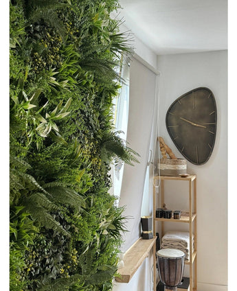 Een weelderige Kunsthaag Freshness 100x100 cm UV verticale tuin bedekt één muur van een lichte kamer. Vlakbij hangt een moderne driehoekige wandklok boven een houten plank met handdoeken, een djembé trommel, manden en kleine decoratieve stukken.