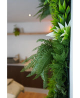 Un primer plano de exuberantes plantas artificiales UV Green Hedge Freshness de 100x100 cm adorna una pared interior vertical, con un fondo borroso que muestra muebles neutros y una pequeña planta colgante.