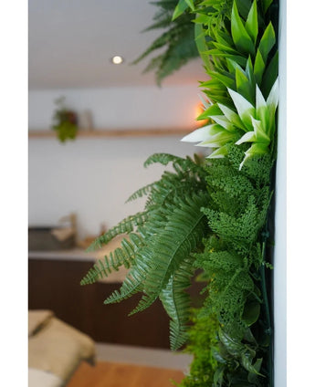 Un primer plano de exuberantes plantas artificiales UV Green Hedge Freshness de 100x100 cm adorna una pared interior vertical, con un fondo borroso que muestra muebles neutros y una pequeña planta colgante.