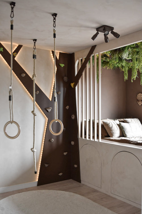Una moderna sala de juegos infantil cuenta con un rocódromo, anillas de gimnasia, cuerdas y un acogedor rincón de lectura con cojines y listones verticales. El seto artificial Lush Deco Deluxe 100x100 cm UV añade vegetación al techo y complementa los tonos neutros y la iluminación divertida de la habitación.