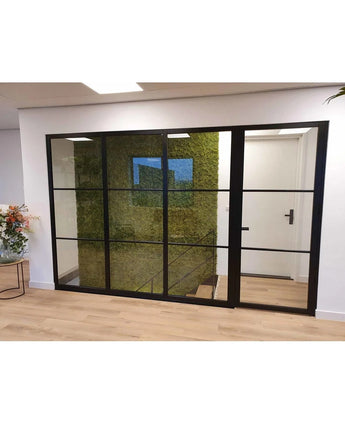 Een modern luxe interieur met een zwarte glazen wand onthult een groene wand van Kunsthaag Nordic Mos 50x50 cm UV, een trap erachter, lichte houten vloeren en een stijlvol bloemstuk aan de linkerkant.