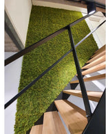 Een moderne trap met houten treden en zwarte leuningen staat naast een groene muur van Kunsthaag Nordic Mos 50x50 cm UV en voegt een natuurlijk, gestructureerd accent toe aan het luxe interieur.